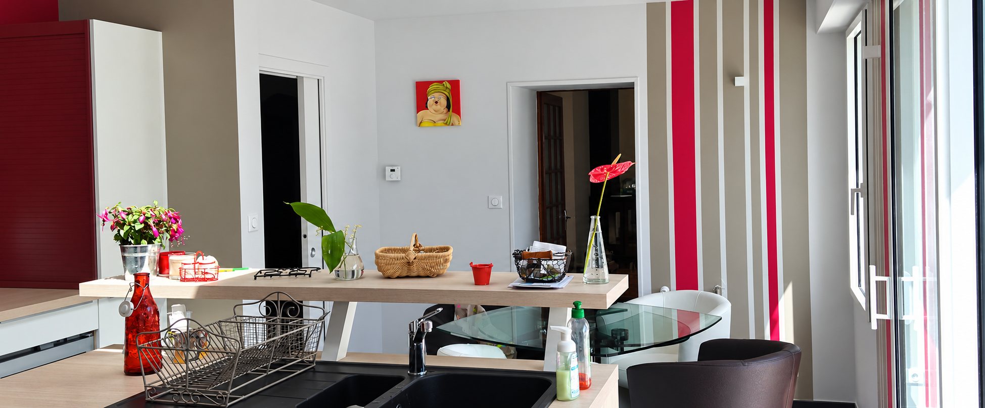 Intérieur d'une cuisine ouverte avec évier, bar en bois clair décoré de fleurs et mur rayé rouge et beige près d'une grande baie vitrée.
