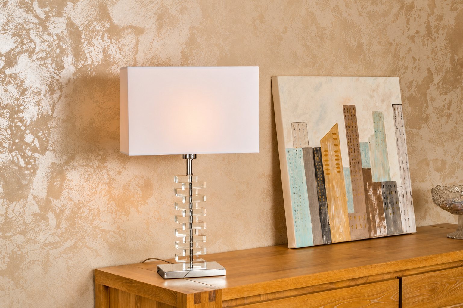 Une lampe de table à abat-jour rectangulaire blanc et pied en cristal est posée sur un buffet en bois clair, à côté d'un tableau de gratte-ciel, devant un mur patiné beige et doré.